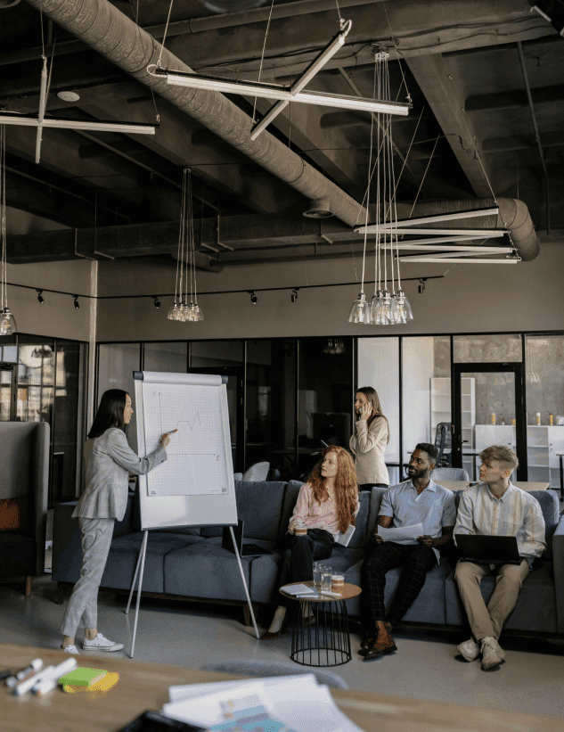 Kobieta prowadząca prezentację na temat wzrostu biznesu, przedstawiająca wykres na tablicy, podczas gdy zespół słucha i notuje.